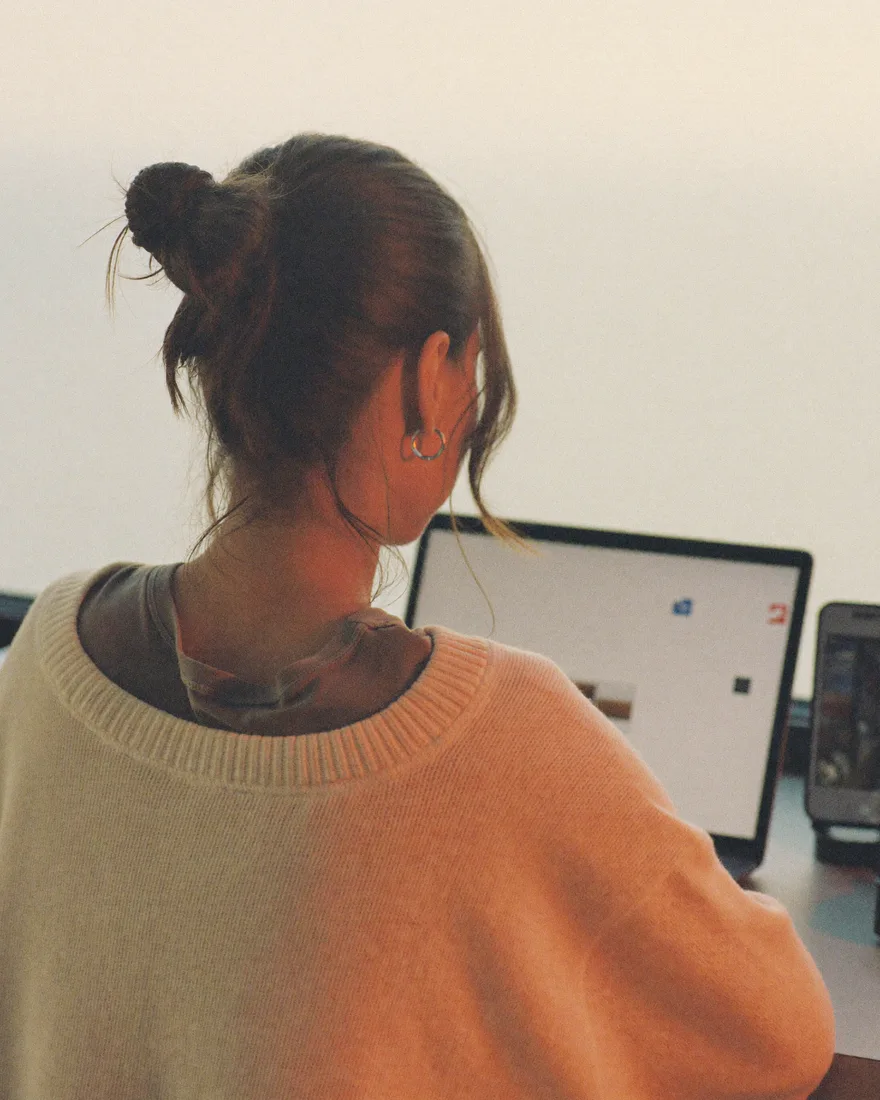 A woman at her desk in the afternoon, seen from behind, facing an open laptop with her phone on a small stand beside it