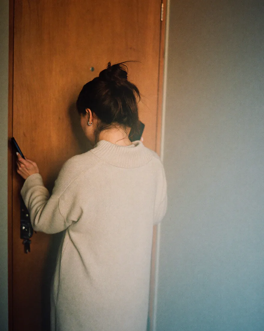 A woman in a long cream sweater at her apartment door in warm hallway light, seen from behind, keys in her hand and a phone at her ear