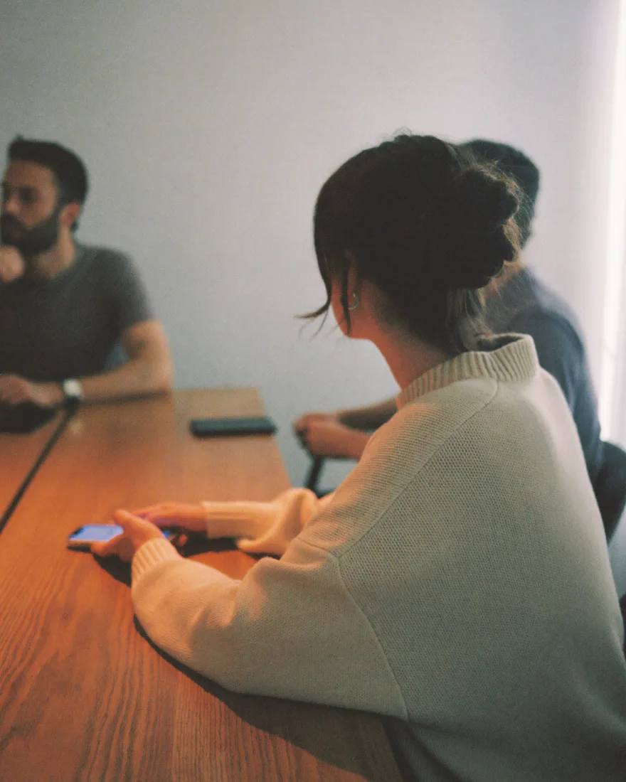 A woman in a cream sweater at a long wooden meeting table, two colleagues sitting nearby