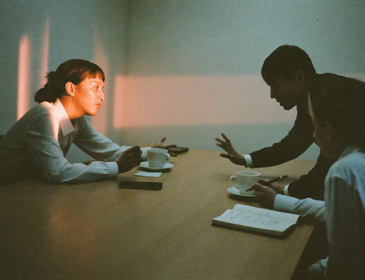 Three colleagues talking around a table in late-afternoon light, a phone lying between their coffee cups