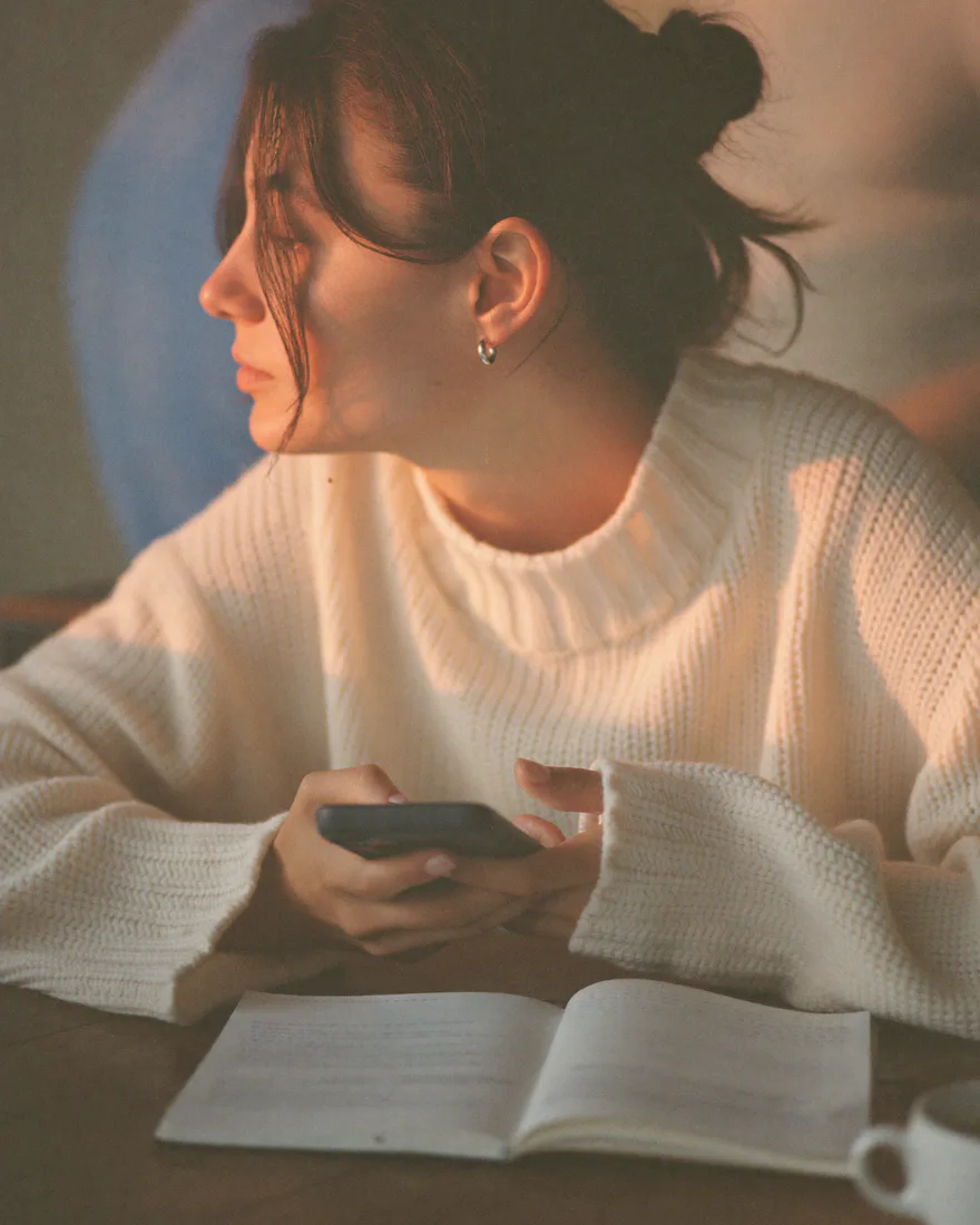 A woman in a cream sweater at a wooden table, holding her phone over an open notebook beside a coffee cup