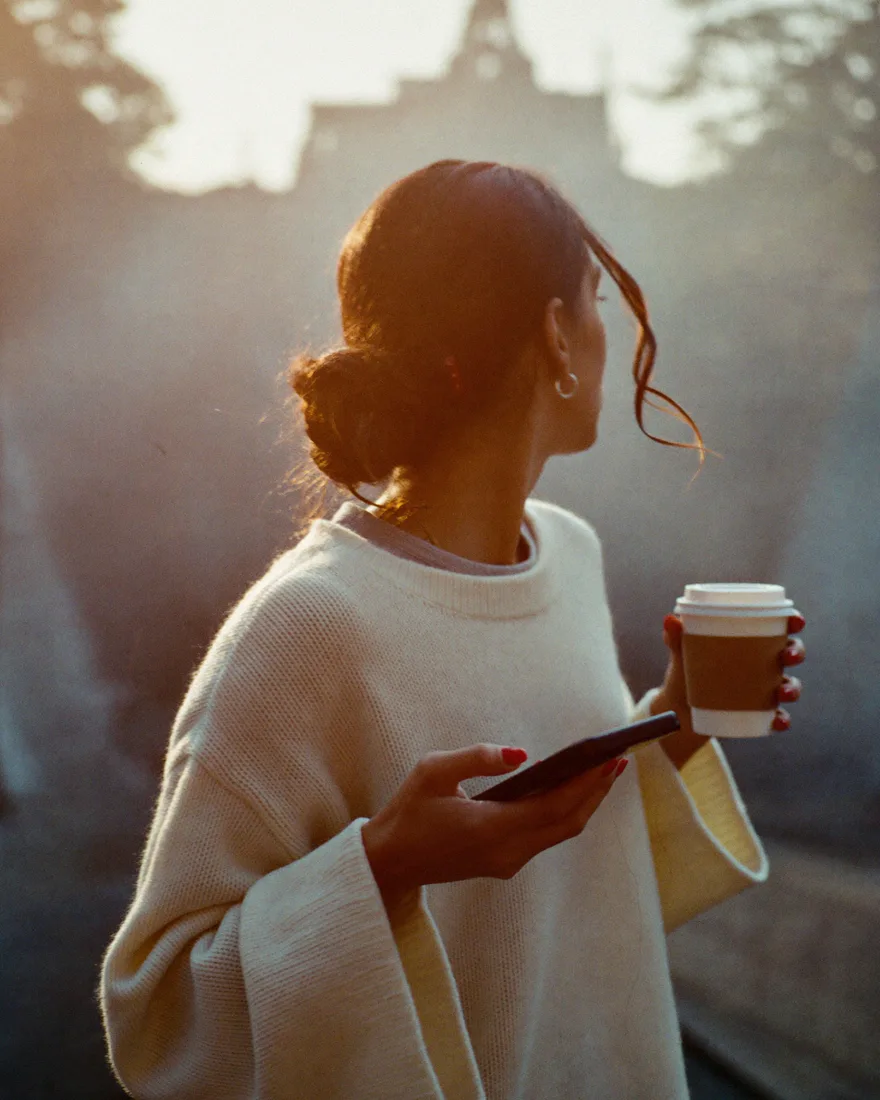 A woman in a cream sweater on a hazy, tree-lined street in the early morning, a phone in one hand and a takeaway coffee in the other