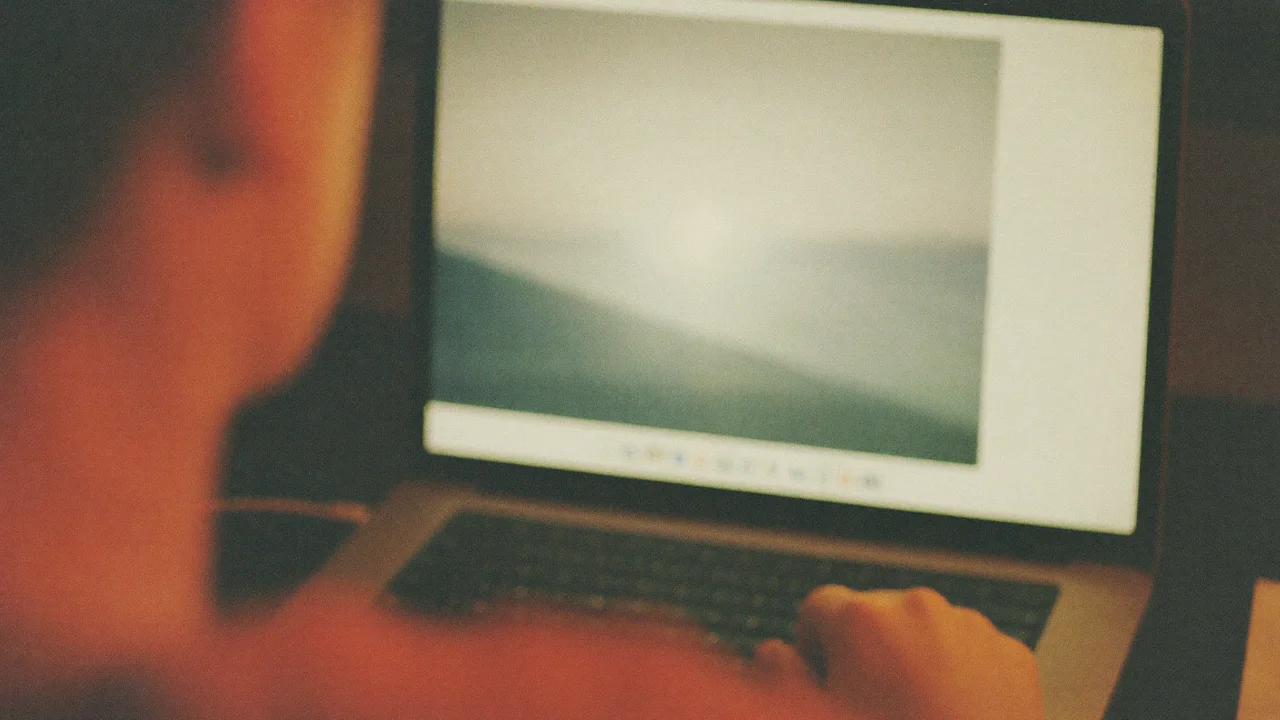 Over the blurred shoulder and cheek of a person, an open laptop shows a soft landscape on its screen while their hand rests on the keyboard, in warm dim light.
