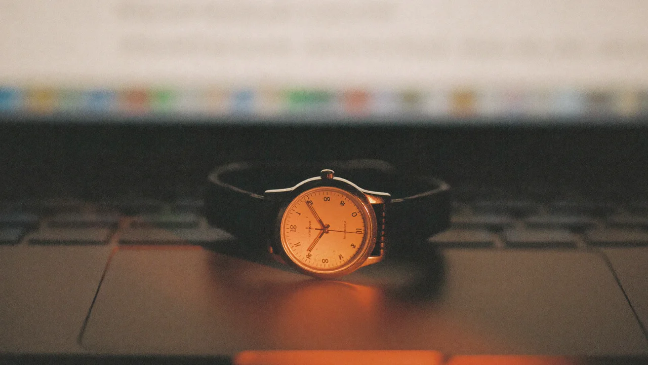 A vintage wristwatch with a cream dial and a dark leather strap lies on a laptop's trackpad in a low warm glow, with the blurred keyboard and screen behind it.