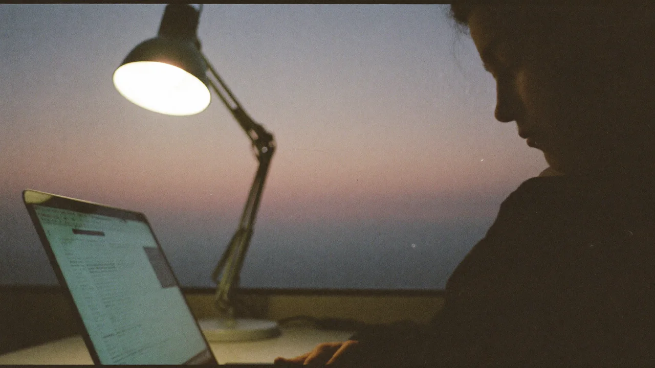 A person in deep shadow sits at a desk at dusk looking down at the glowing screen of an open laptop, beside an angled desk lamp, with a pink and blue twilight sky through the window.