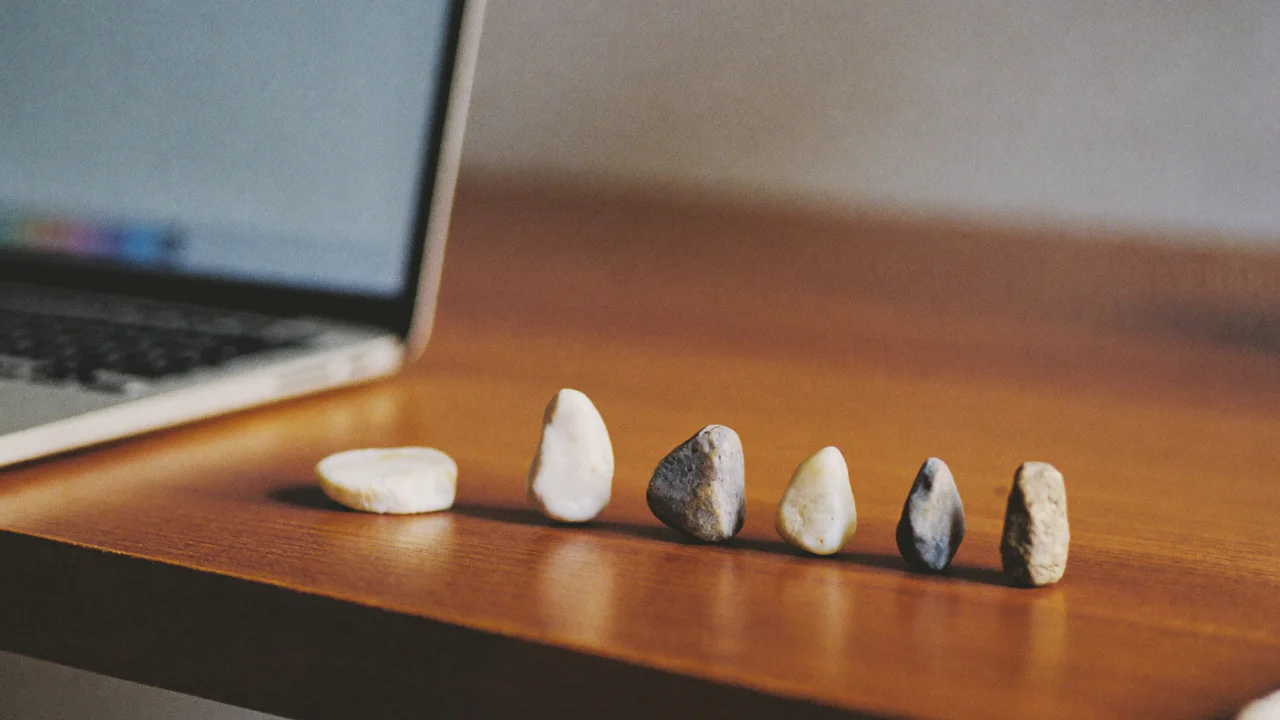 Six small smooth stones in white, grey and tan stand in a row on a warm wooden desk beside an open laptop, its screen out of focus.
